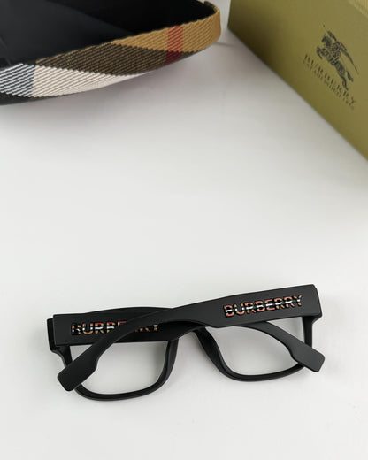 Black Burberry glasses on a white surface with a checkered hat and yellow box in the background.